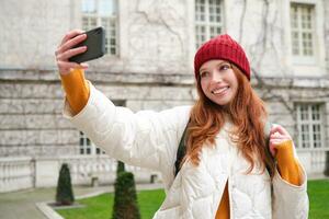 Cute ginger girl in red hat takes selfie during her tourist trip abroad. Young redhead woman makes a photo of herself in front of historical landmark