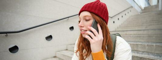 joven elegante pelirrojo niña en rojo sombrero, se sienta en calle y negociaciones en móvil teléfono, tiene teléfono conversación, anillos su amigo mientras relaja al aire libre foto