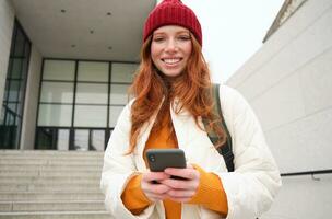 Happy girl student in red hat, holds smartphone, tourist looks at map app on her phone, explores sightseeing, texts message, looks for couchsurfing, rents place to stay online photo