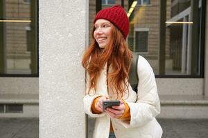 Beautiful redhead girl with smartphone, standing on street, holding mobile phone, using taxi app, connects to public wifi and uses internet, sends a text message photo
