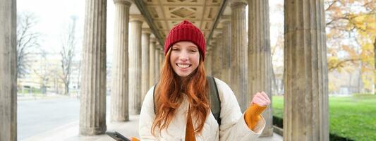 Mobile broadband and people. Smiling redhead 20s girl with backpack, uses smartphone on street, holds mobile phone and looks at application photo