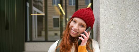 Happy beautiful girl, redhead tourist talks on mobile phone, making a call abroad, has conversation on smartphone, standing with backpack outdoors photo
