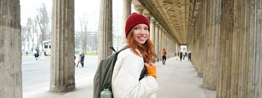 Female tourist with red long hair, holds backpack with thermos, turns back at camera with happy smile, explores city and popular tourism landmarks photo