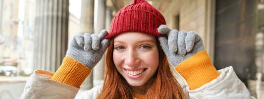 Headshot of happy redhead girl with freckles, wears red hat and gloves in winter, walks around city on chilly weather and smiles photo