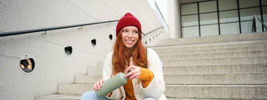 Smiling traveler, redhead girl tourist sits on stairs with flask, drinks hot coffee from thermos while travelling and sightseeing around foreign city, sits on stairs and rests photo