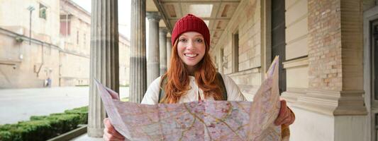 sonriente joven pelirrojo mujer en rojo sombrero, mira a papel mapa a Mira para turista atracción. turismo y personas concepto. niña explora ciudad, intentó a encontrar camino foto