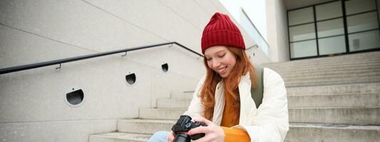 Smiling redhead girl photographer, checks her shots, holds camera and looks at screen, takes photos outdoors, walks around street and does streetstyle shooting