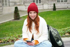 Young smiling redhead girl sits in park with smartphone, texting message on mobile phone, using telephone, connecting to public wifi and surfing internet photo