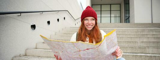 hermosa niña turista se sienta en escalera con ciudad mapa, planes su viaje, mira para dirección mientras de viaje alrededor ciudad, búsquedas ruta para Turismo foto