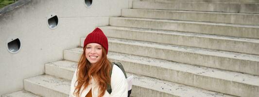 Smiling redhead girl, student sits on stairs outdoors and uses laptop, connects to public wifi in city and works on project, uses internet on computer photo