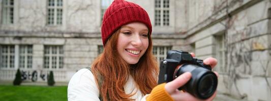 Redhead girl photographer takes photos on professional camera outdoors, captures streetstyle shots, looks excited while taking pictures