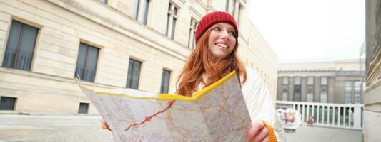 pelirrojo chica, turista explora ciudad, mira a papel mapa a encontrar camino para histórico puntos de referencia, mujer en su viaje alrededor Europa búsquedas para Turismo foto