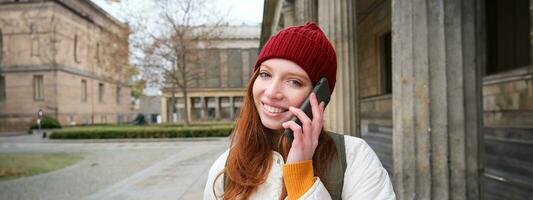 sonriente joven pelirrojo mujer escucha a voz mensaje, hace un teléfono llamar, camina en calle y negociaciones a alguien en teléfono inteligente foto