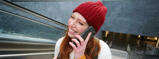 Portrait of happy redhead woman walking around town with smartphone, calling someone, talking on mobile phone outdoors photo