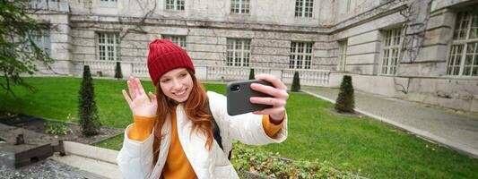 Portrait of girl blogger records video and says hi at phone camera. Young woman takes selfie with smartphone app, waves hand, video chats in application photo