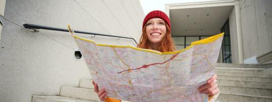 joven sonriente pelirrojo chica, turista se sienta en escalera al aire libre con ciudad papel mapa, mirando para dirección, viajero mochilero explora ciudad y mira para Turismo foto