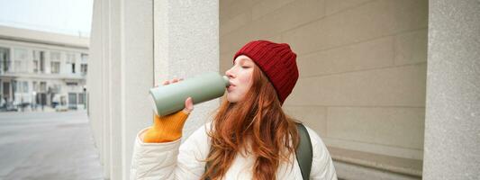 Beautiful redhead woman drinking hot tea or coffee from thermos, female tourist enjoys warm drink, rests during her journey around town photo