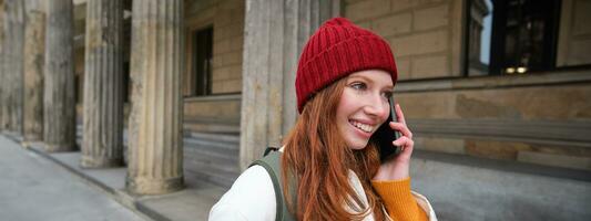 sonriente joven pelirrojo mujer escucha a voz mensaje, hace un teléfono llamar, camina en calle y negociaciones a alguien en teléfono inteligente foto