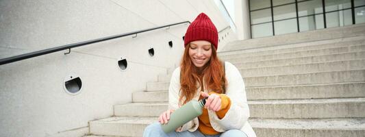 Happy redhead woman, tourist with thermos, drinks her hot tea, coffee from travel flask, restests during her travelling in city and sightseeing photo