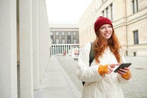 Beautiful smiling girl, tourist with backpack, holding smartphone, using map on mobile phone application, looking for sightseeing in internet app, standing outdoors photo