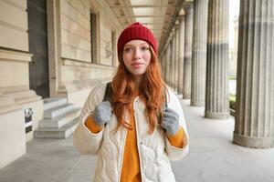 Smiling redhead girl with backpack, walks in city and does sightseeing, explore popular landmarks on her tourist journey around Europe photo