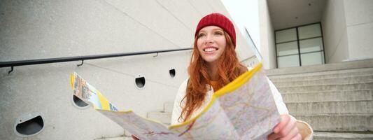 joven sonriente pelirrojo chica, turista se sienta en escalera al aire libre con ciudad papel mapa, mirando para dirección, viajero mochilero explora ciudad y mira para Turismo foto