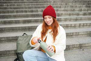Smiling tourist, girl sits on stairs, rests on staircase, takes thermos from backpack, drinks hot coffee from flask photo