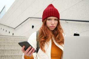 Portrait of redhead woman sits on stairs, uses laptop and holds smartphone, looks confused and upset at computer screen, tries to connect to wifi public internet photo
