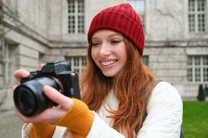 Redhead girl photographer takes photos on professional camera outdoors, captures streetstyle shots, looks excited while taking pictures