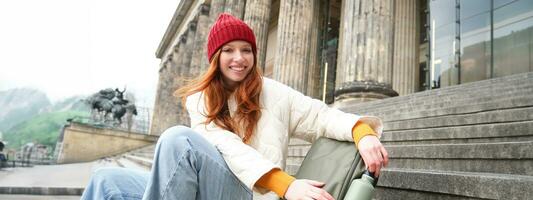 Young redhead female tourist rests during her trip, opens thermos and drinks hot tea, having a break after sightseeing photo