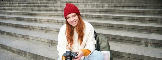 retrato de joven fotógrafo chica, se sienta en escalera con profesional cámara, toma fotos al aire libre, haciendo estilo de vida disparo