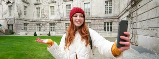 Portrait of happy tourist, girl with backpack, video chats, shows building, sightseeing attraction to friend, poses near landmark and smiles photo