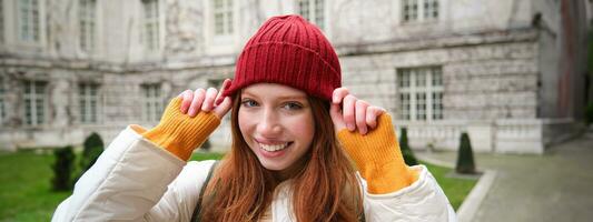 Young smiling redhead woman walking in beautiful city attractions, wearing backpack, red hat and coat, looking around with happy face photo