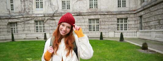 Beautiful redhead woman with backpack, tourist walking around city park and looks around, wears red hat and warm coat photo