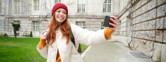 Portrait of happy girl tourist, takes selfie on smartphone in front of historical building, posing for photo on mobile phone camera