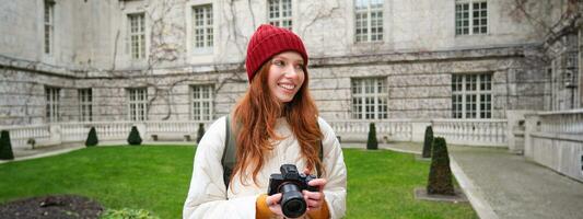 Redhead girl photographer takes photos on professional camera outdoors, captures streetstyle shots, looks excited while taking pictures