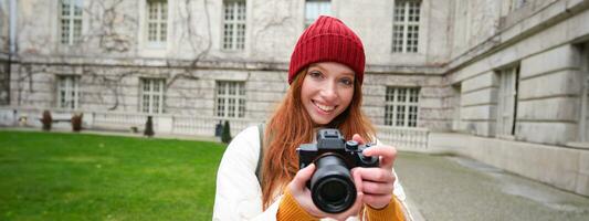 Redhead girl photographer takes photos on professional camera outdoors, captures streetstyle shots, looks excited while taking pictures