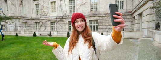 Portrait of happy girl, tourist posing near city attraction, video chats and demonstrates landmark, shows something to friend with mobile phone app photo