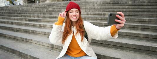 Stylish young girl in red hat, takes photos on smartphone camera, makes selfie as she sits on stairs near museum, posing for photo with app filter