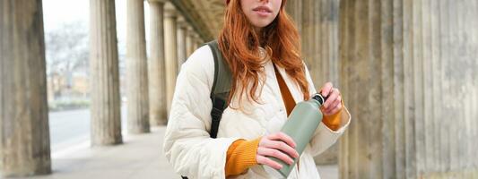 Close up portrait of redhead girl, tourist stands on street, opens thermos with hot drink, rests and pours herself coffee from flask photo