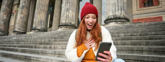 pelirrojo hembra turista negociaciones en móvil teléfono, vídeo chateando con amigos, dice Hola y olas mano a teléfono inteligente cámara foto