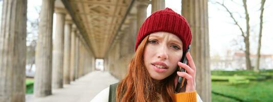 Portrait of redhead girl in red hat, calls someone, listens to voice message with concerned, confused face expression, using smartphone photo