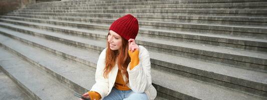 Portrait of young urban girl in red hat, sits on stairs near museum, holds mobile phone, connects to public wifi and surfs net, uses smartphone apps photo