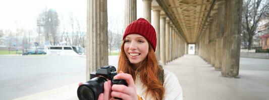 Young redhead female photographer, makes lifestyle shooting in city centre, takes photos and smiles, looks for perfect shot, makes picture