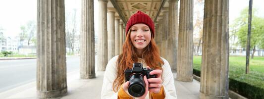 Smiling tourist photographer, takes picture during her trip, holds professional camera and makes photos