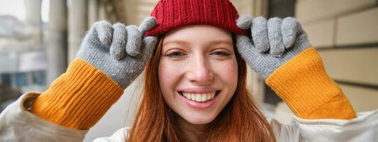 disparo a la cabeza de contento pelirrojo niña con pecas, usa rojo sombrero y guantes en invierno, camina alrededor ciudad en frío clima y sonrisas foto