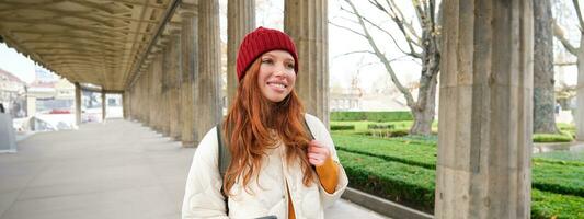 Mobile broadband and people. Smiling redhead 20s girl with backpack, uses smartphone on street, holds mobile phone and looks at application photo