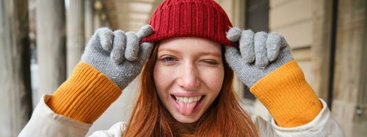 Portrait of young redhead woman in knitted hat and gloves, smiles and looks aside, walks around city in winter photo
