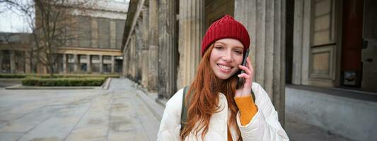 Smiling redhead female tourist talks on mobile phone and walks around city. Happy student in red hat calls friend, stands on street and uses smartphone photo