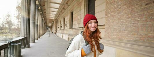 Smiling redhead girl with backpack, walks in city and does sightseeing, explore popular landmarks on her tourist journey around Europe photo